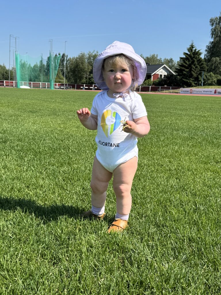 Kuvassa noin vuoden ikäinen lapsi seisoo urheilukentällä. Päällään hänellä on Kuortaneen kunnan logolla varustettu valkoinen body. Päässä valkoinen hattu.