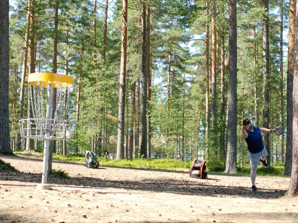 Mies pelaa frisbeegolfia metsässä. Heittää kiekkoa kuvan oikealta kuvan vasemmassa olevaan koriin.