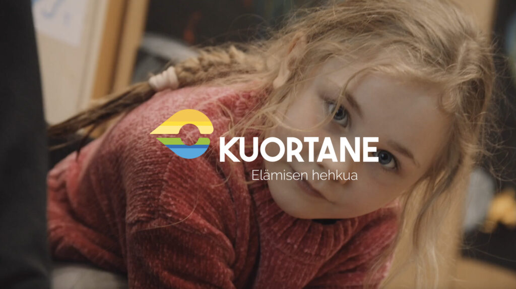 Tyttö punaisessa paidassa istuu eteenpäin kumartuneena ja katsoo kameraan.
Kuvan päällä on Kuortaneen kunnan logo.