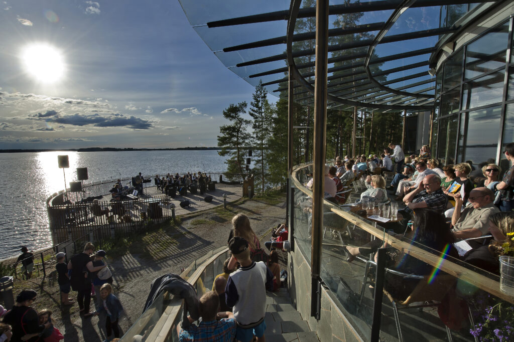 Kuortaneen Urheiluopiston terassi kesäisen aurinkoisena päivänä. 
Terassilla paljon ihmisiä, Kuortaneenjärven edessä olevalla lavalla sooittaa puhallinorkesteri.