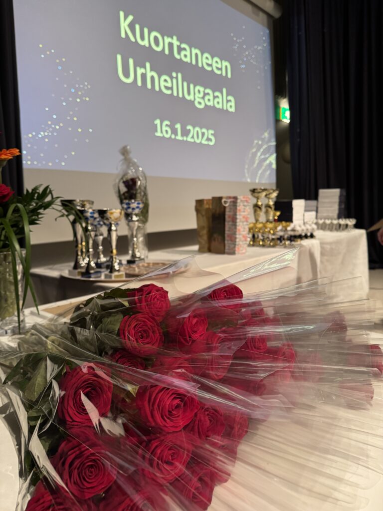 Ruusuja ja pokaaleja pöydällä. Taustalla kankaalle heijastettu teksti Kuortaneen Urheilugaala.