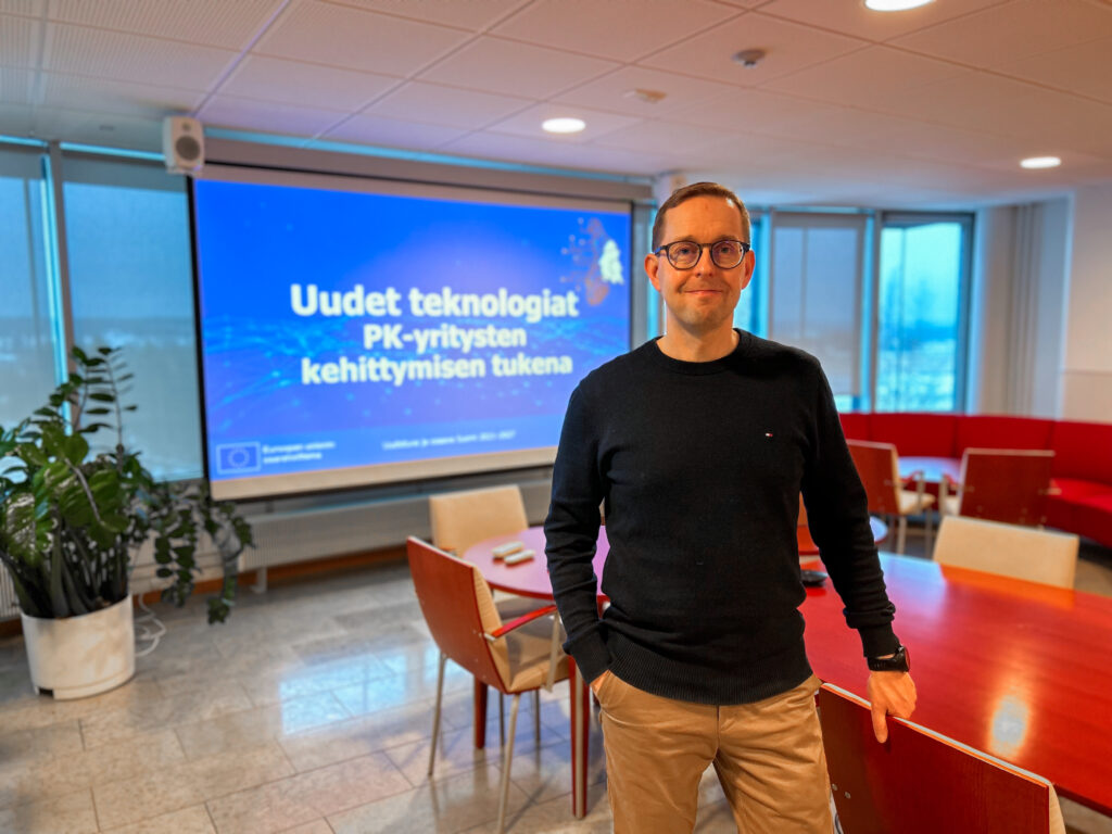 Kuvassa oikealla projektipäällikkö Hannu Ylinen seisoo punaisen pöydän edessä. Vasemmalla takana screenseinä, jossa teksti: Uudet teknologiat pk-yritysten kehittymisen tukena.