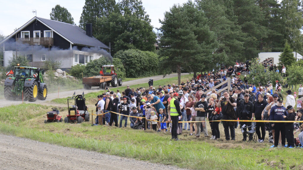 Traktorien mäkivetokisat. Kaksi traktoria kuvan vasemmalla kulkevat mäkeä ylöspäin. Kuvan oikealla puoella on satapäinen väkijoukko seuraamassa.
