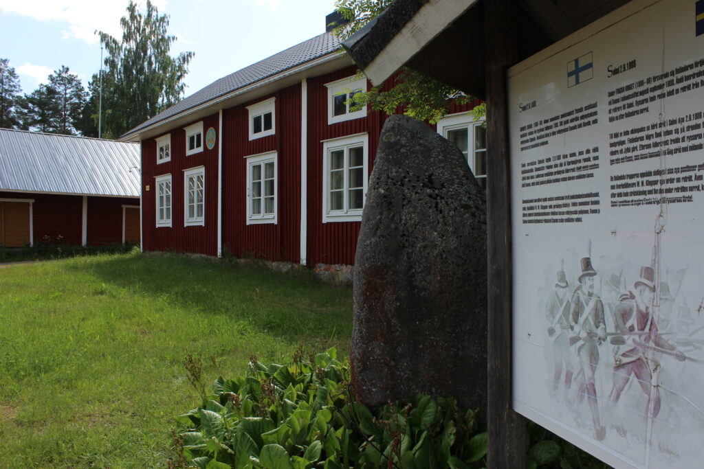 Punainen pohjalaistalo Salmen kylällä, jonka vieressä vuoden 1918 vapaussotaan lähdöstä kertova infokyltti.